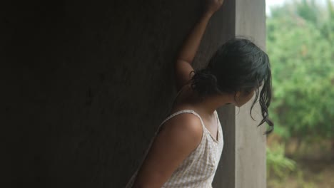 una mujer con un vestido blanco se encuentra pensativa junto a una ventana en un entorno rústico, mirando hacia afuera