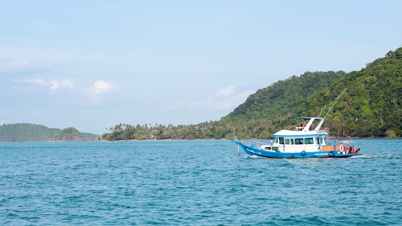 A boat travels across clear blue waters near lush green hills in Phuket, Thailand, under bright daylight