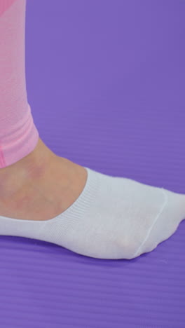 primer plano de una pierna estirada en una alfombra de yoga con calcetines blancos y leggings rosados, participando en un entrenamiento, vista parcial de la mano balanceándose en movimiento, con un fondo púrpura suave