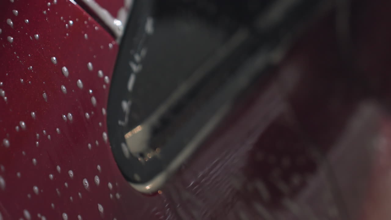 Macro view of soap dripping down glossy red car surface during washing process showing texture of water mixed with detergent forming streaks and bubbles on bodywork inside garage environment
