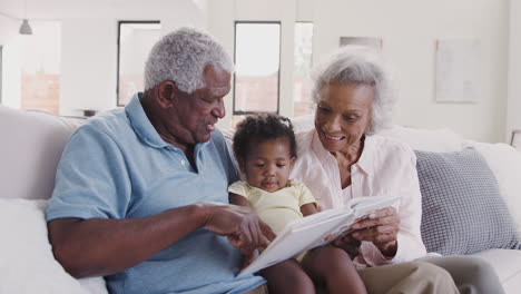 grandparents sitting on sofa at home reading book with baby granddaughter