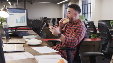 happy biracial casual businessman sitting at desk, eating sandwich and using smartphone, slow motion