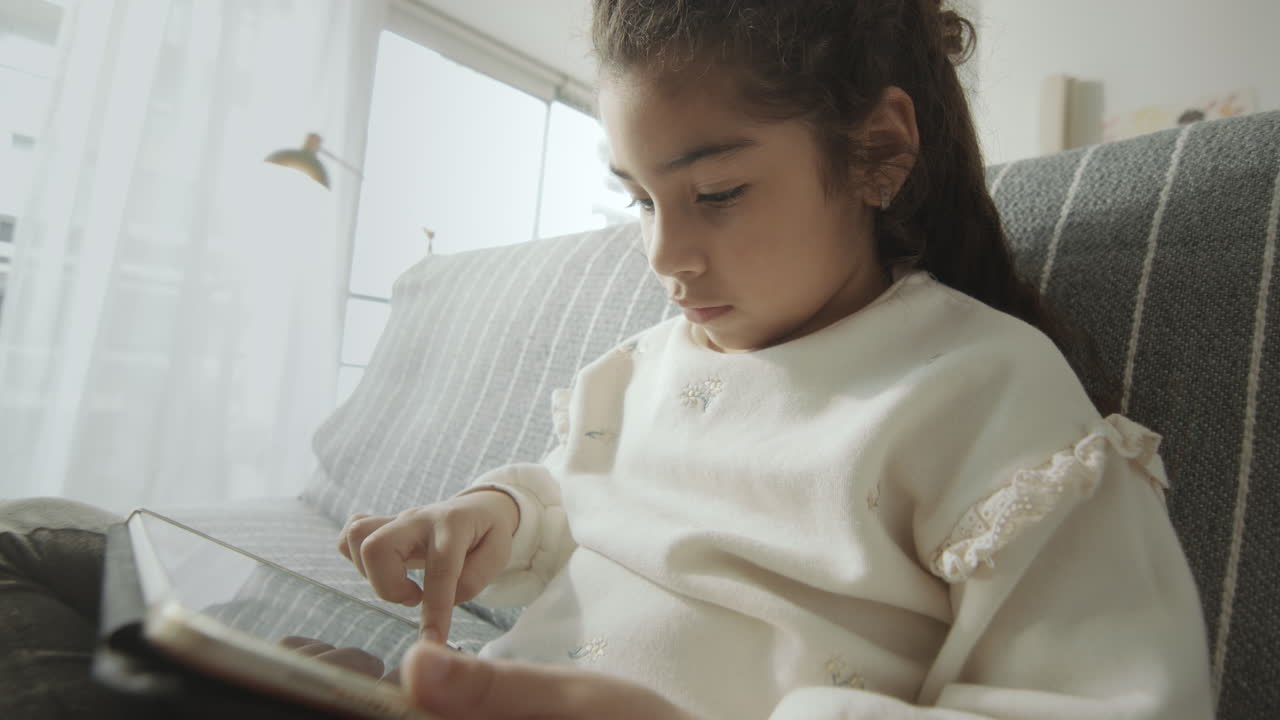 Pretty Little Girl Sitting on Sofa and Using Digital Tablet