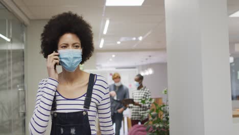 Portrait-of-african-american-creative-businesswoman-wearing-face-mask-using-smartphone-in-office