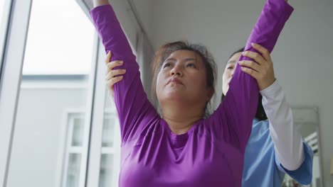 Asian-female-physiotherapist-helping-female-patient-exercise-arms-with-exercise-band-at-surgery