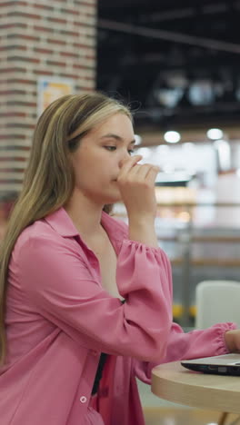 dama blanca sentada sola en un restaurante ocupado operando su portátil, se inclina hacia adelante, toma una respiración profunda, y mirando cansada coloca su mano debajo de su barbilla, la mujer en el fondo está detrás