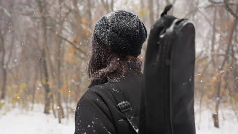 side view of lady in black beanie and hoodie, speckled with snow, walking through a snow-covered park with a guitar pack, surrounded by bare, frost-laden trees