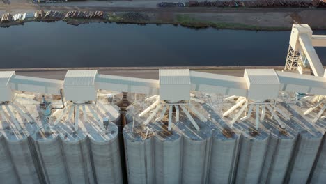 aerial top down view tall grain storage silo facility bordered by rail road tracks in rail yard with train cars parked on waterfront - drone flyover