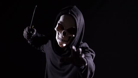 close up of scary man in the hooded sweatshirt wearing halloween mask holding a knife and making frightening gesture while running on the black background