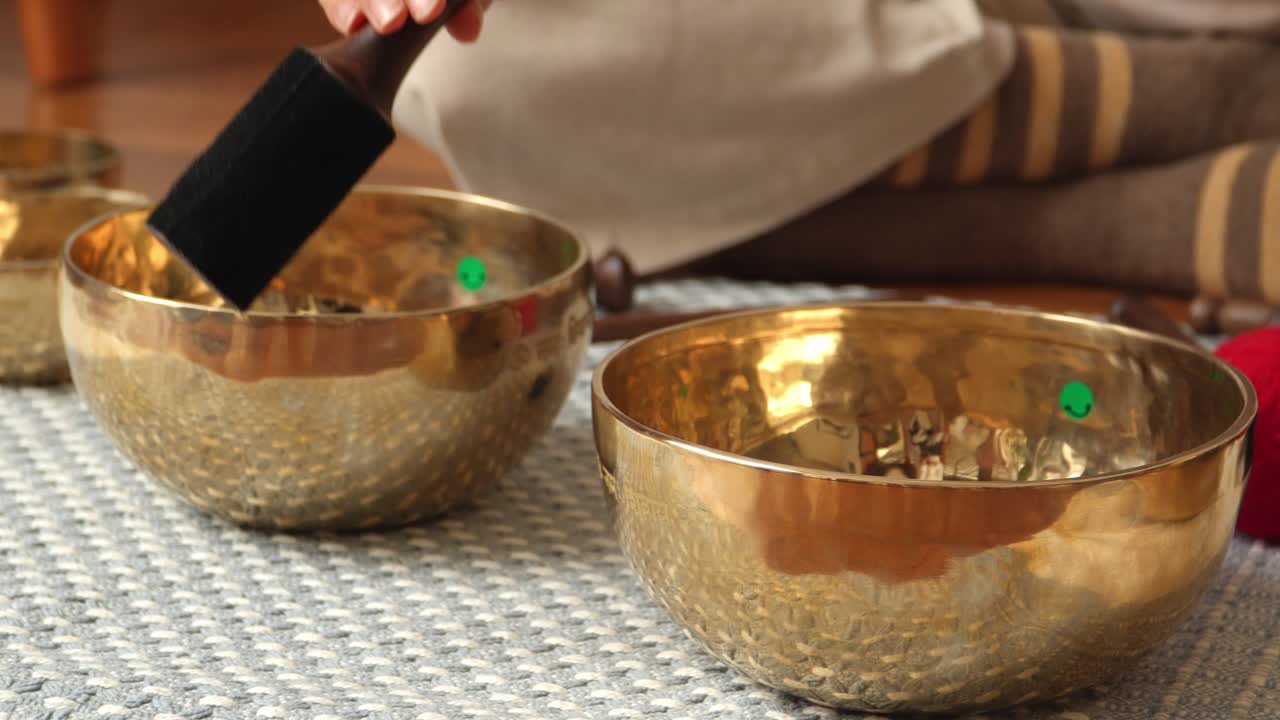 Dynamic close-up moving shot as a hand strikes and circles several brass Tibetan singing bowls arranged on a woven mat in a calm indoor wellness studio