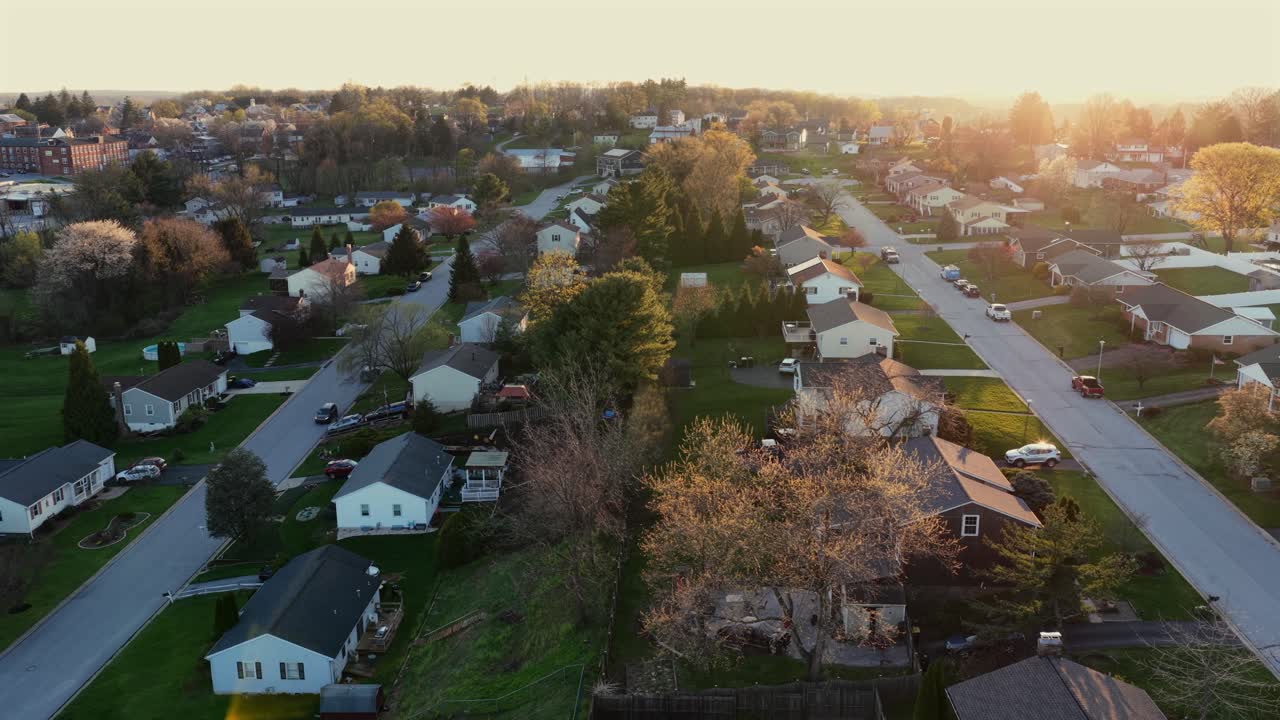 Middle-class American suburban neighborhood at sunset, with warm golden light, spring trees in bloom, quiet residential streets and single-family homes in Pennsylvania. Aerial lateral wide shot
