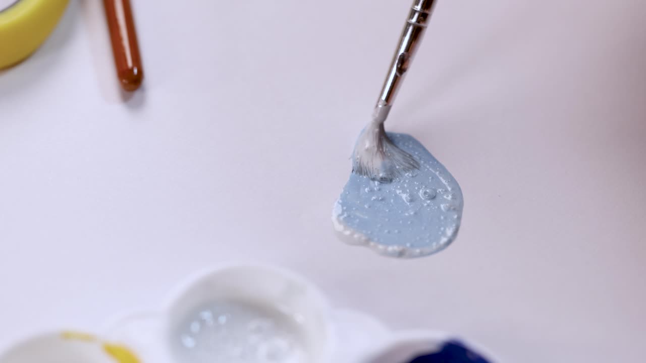Artist mixes light blue paint with brush on white palette under soft, even lighting
