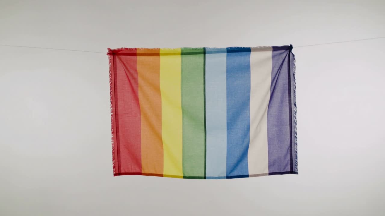 Waving vertical rainbow flag fluttering on thin rope under pale sky, breeze causing folds settling