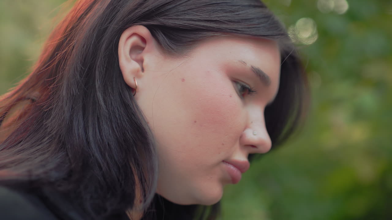 close up side view of fair skinned girl, attention focused on her book reading, Adjusting hair by side of her ear, blur forest background, hair adjusting beside ear, blur forest background