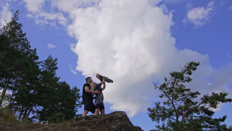 caucasian couple stand on the rock. woman holding her baby over the head. young family resting in the nature. low angle view.
