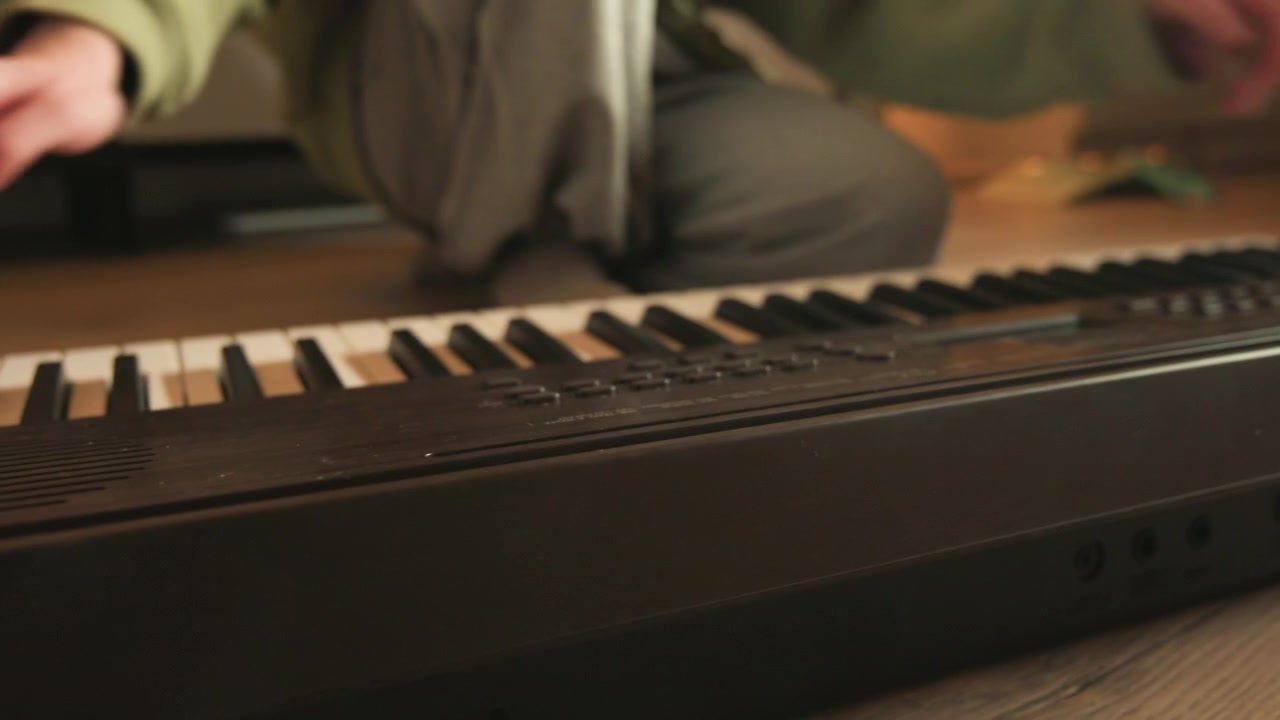 Närbild i låg vinkel av ett elektroniskt keyboard med händer som trycker på tangenter och fötter på ett trägolv, som fångar synlig spelrörelse och subtila viktförskjutningar under avslappnad hemövning.