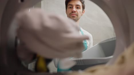 View-from-inside-the-washing-machine-a-confident-brunette-guy-in-a-white-T-shirt-and-a-blue-apron-throws-dirty-things-into-the-washing-machine-and-closes-it