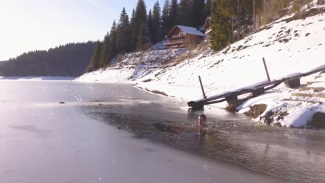 Drone-flying-low-over-ice-covered-lake-surface-with-man-sitting-in-water-up-to-face