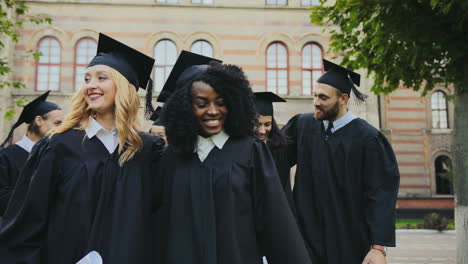 glücklich lächelnde multiethnische absolventen, die nach der abschlussfeier mit den diplomen in den händen in der nähe des universitätsgebäudes spazieren