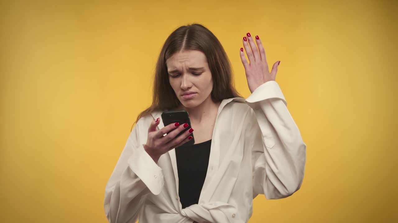 una joven caucásica adulta está molesta por las noticias en su teléfono inteligente sobre un fondo amarillo brillante
