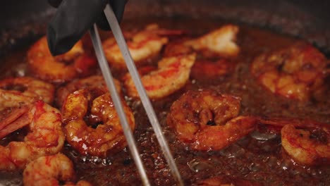 hand of a professional chef using stainless steel tongs, flipping sides of delicious peeled prawns ensuring the shrimps are cooking evenly in tasty bubbling red sauce, extreme close up shot