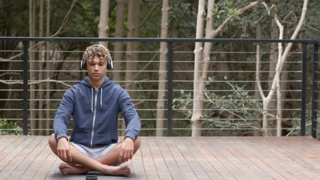 young man meditating outdoors with headphones, relaxing on wooden deck