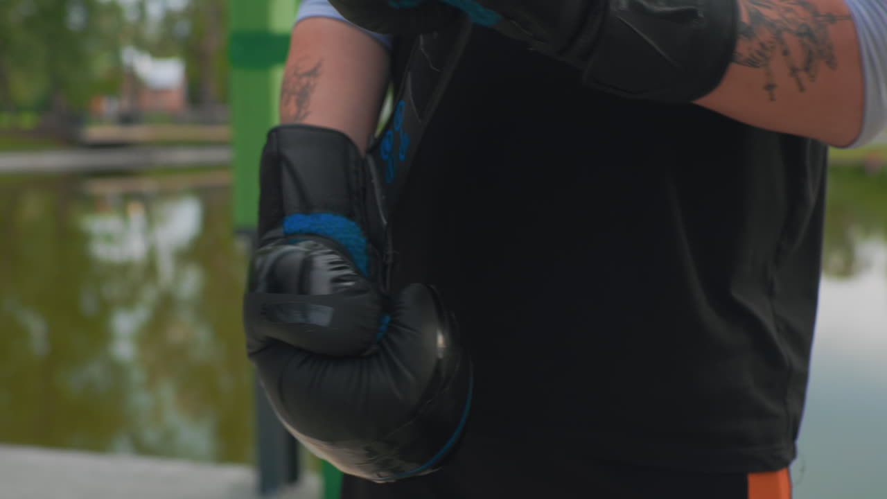 white man tattooed forearm adjusting boxing gloves in closeup, detailed leather straps and mitts, trainer or fighter tightening grip, raw texture and sweat, lakeside backdrop, intense preparation