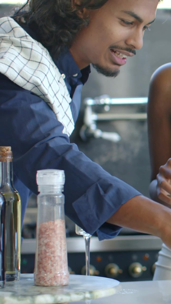 Vertical video: Smiling man with towel giving item across kitchen counter to her, with grinder