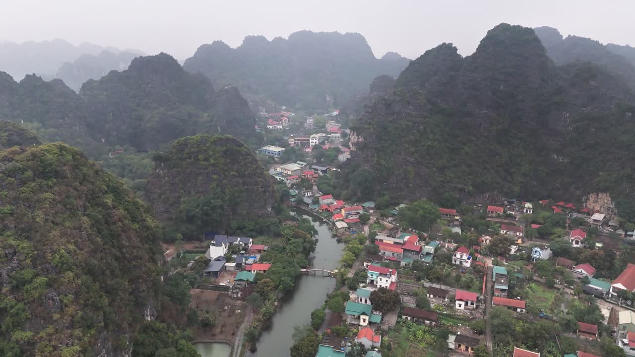 Aerial view of Trang An in Ninh Binh, Vietnam, featuring a small village nestled between limestone mountains, lush greenery, and winding waterways under an overcast sky