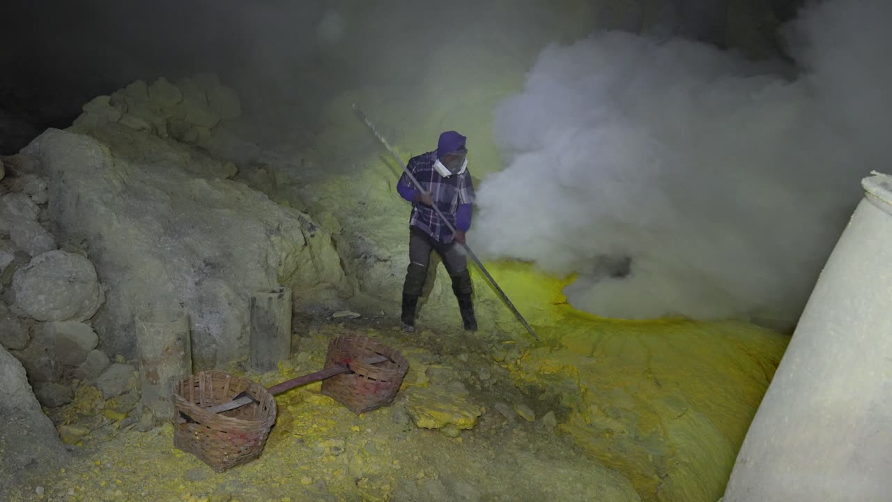 Wide shot shows a masked miner in Kawah Ijen crater using a long pole to break cooling sulfur near a steaming vent, with traditional woven baskets ready for the heavy load