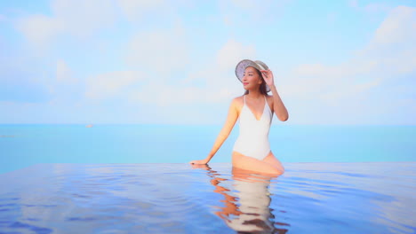 원피스 수영복을 입은 예쁜 젊은 여성이 바다를 배경으로 인피니티 풀 가장자리에 앉아 있습니다.