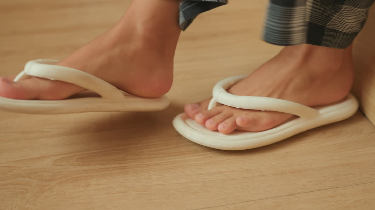 Een hotelgast glipt 's ochtends in pantoffels naast het bed; pyjamavoeten schuiven in zachte schuimsandalen op een houten vloer. Een close-up van de teenbeweging, de linnen rand en warm daglicht roepen een ontspannen ontwaken op.
