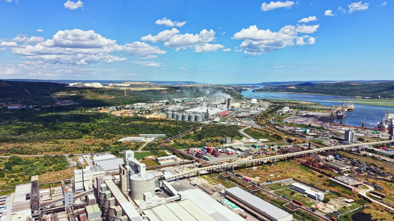 Zona industrial con fábricas, almacenes y un río al fondo en un día soleado con nubes dispersas.