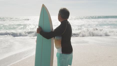 Back-view-of-senior-hispanic-man-standing-on-beach-with-surfboard