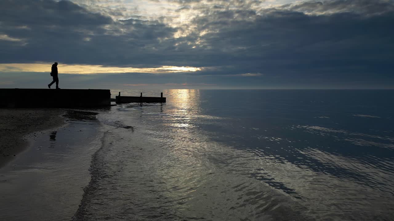 Sakte film av milde bølger med en mann i silhuett som går til enden av moloen ved solnedgang på Fleetwood Beach i Lancashire, Storbritannia.
