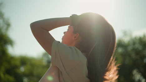 lady in black gloves tilts head back, adjusting her hair in warm sunlight, with serene green trees and foliage softly blurred in background