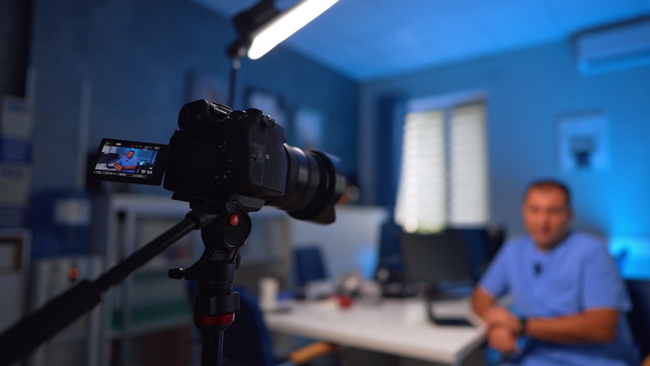 Doctor in blue uniform takes video for a vlog. Medic talks to camera in front of him set on a tripod. Selective focus