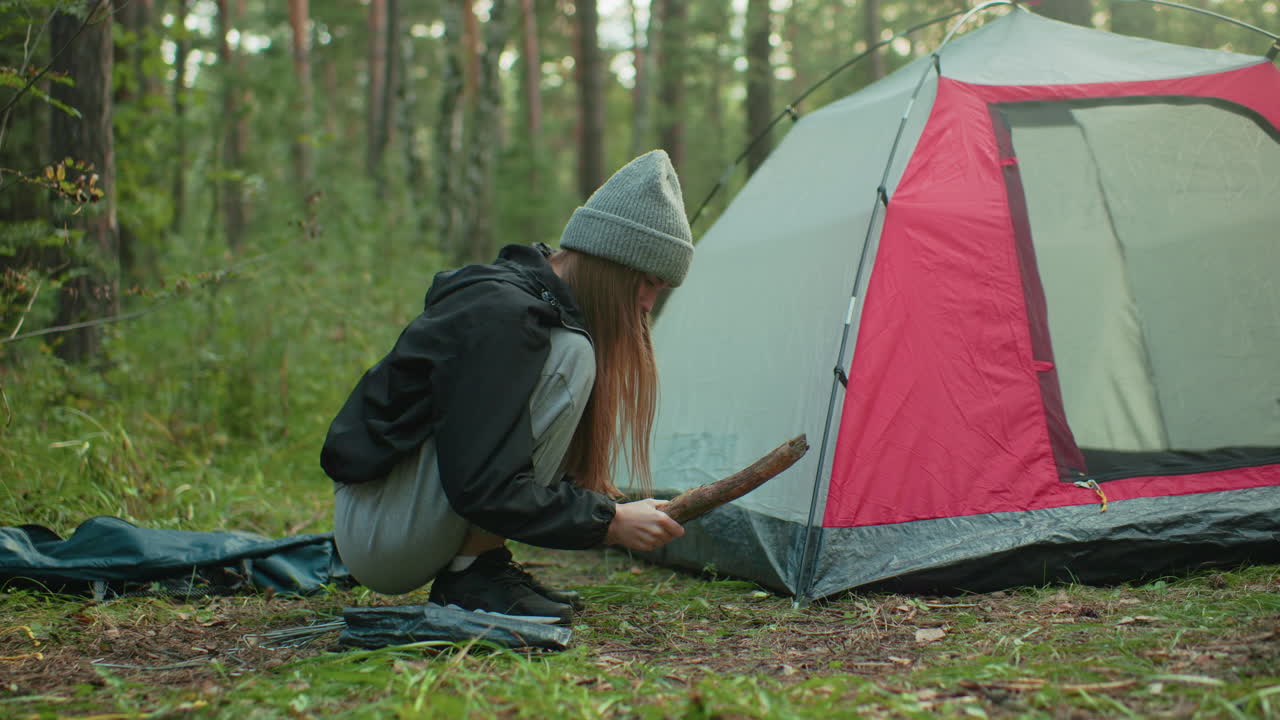 student squats beside camping tent pressing peg into ground while using large stick as hammer surrounded by green forest environment and camping gear during outdoor setup in woodland area