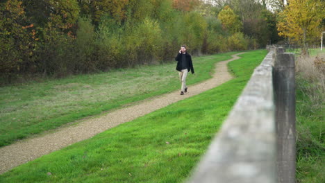 young man on phone call walks down dirt path in a park towards the camera
