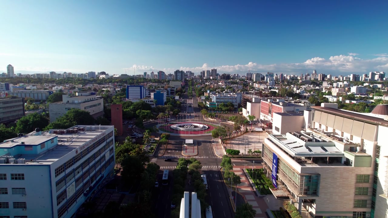 Imagem aérea de drone em ângulo elevado subindo verticalmente sobre o monumento obelisco dos Heróis, passando por uma estátua branca e uma fonte, para revelar a rotatória do Supremo Tribunal e o horizonte da cidade em Santo Domingo.