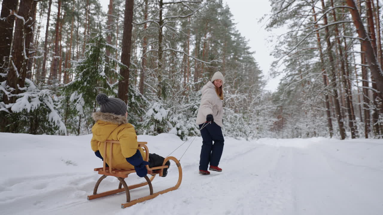 Una giovane madre e suo figlio si divertono in inverno nella foresta andando in slitta al rallentatore. Mamma felice a passeggio con suo figlio in una foresta innevata.