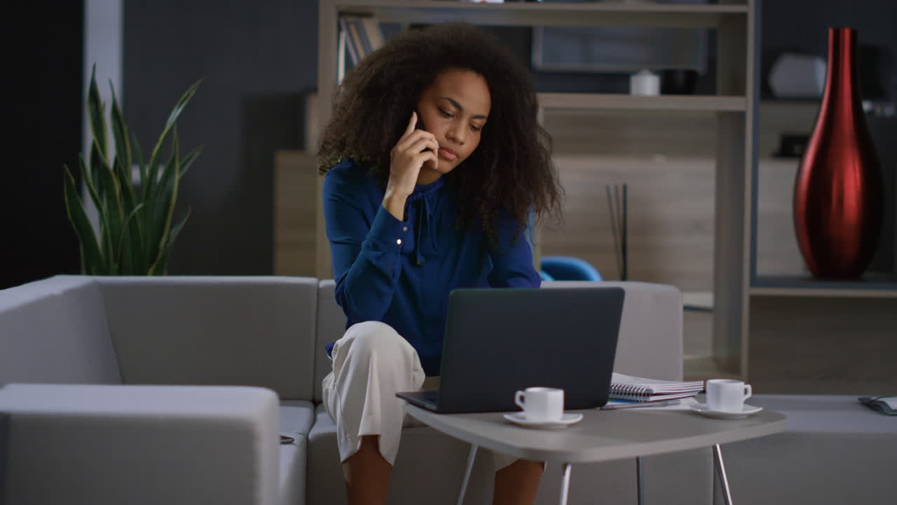 Busy african woman talking mobile phone working laptop computer in remote office