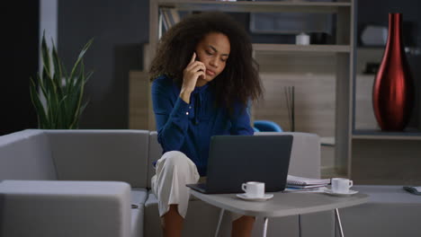 Busy-african-woman-talking-mobile-phone-working-laptop-computer-in-remote-office