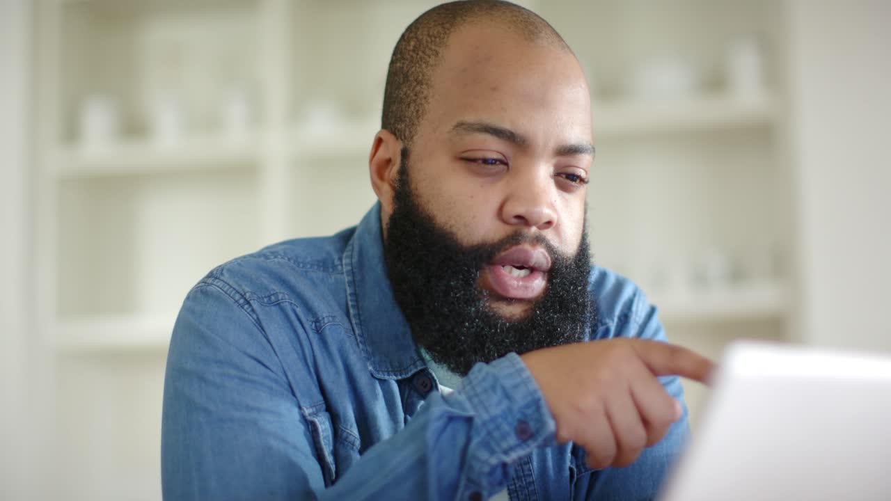 African man looking down at tablet pointing tapping index finger explaining at home in denim shirt