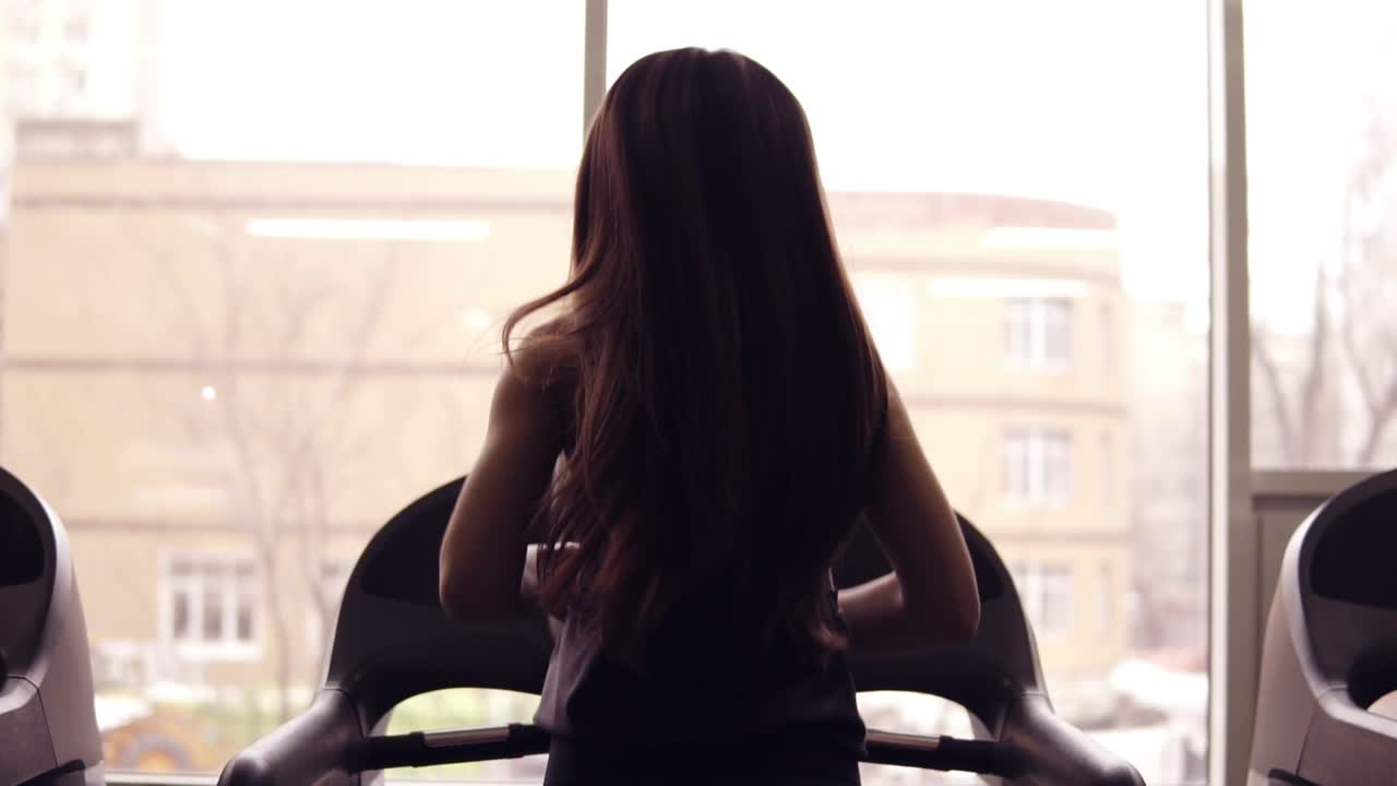 긴 갈색 머리카락을 가진 스포츠 여성 운동 선수가 트레드밀에서 달리는 뒤쪽 영상. 실내. 건강한 생활 방식, 스포츠 및 피트니스, 웰빙