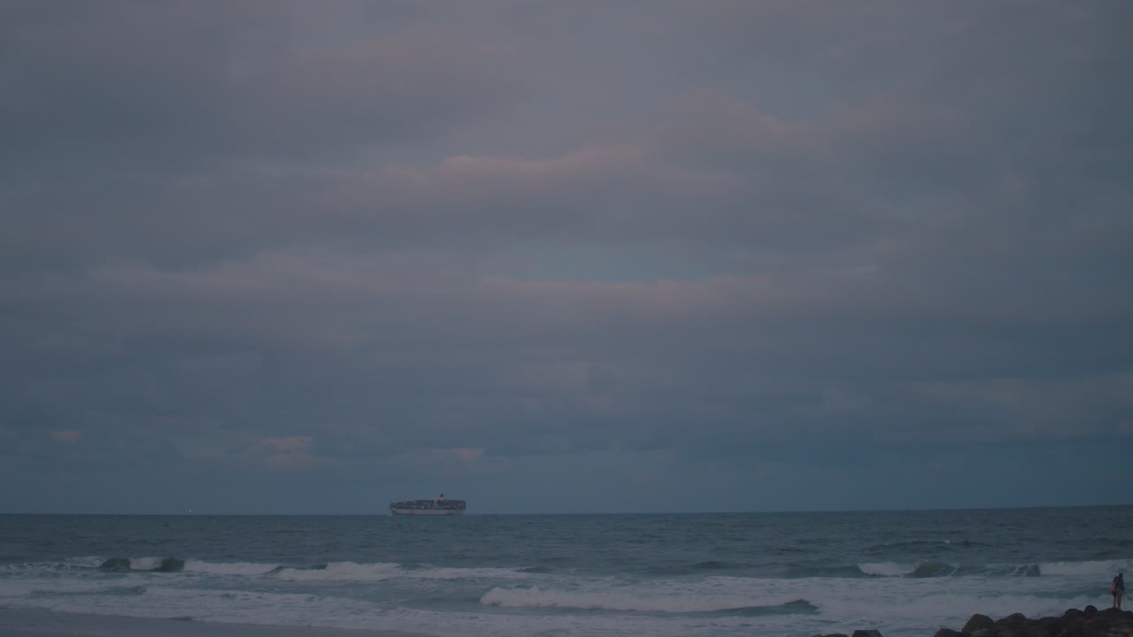석양이 파란색과 분홍색의 밝은 색을 위험하고 폭풍우가 치는 하늘과 아래의 어둡고 변덕스러운 물에 쏟는 오후 해변의 수평선을 따라 대형 화물선이 항해합니다.