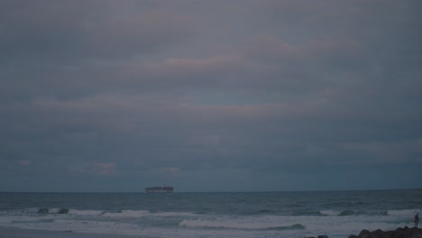 Un-Gran-Barco-De-Carga-Navega-Por-El-Horizonte-De-Una-Playa-De-La-Tarde-Donde-El-Sol-Poniente-Derrama-Colores-Claros-De-Azul-Y-Rosa-En-El-Cielo-Tormentoso-Y-Peligroso,-Así-Como-En-El-Agua-Oscura-Y-Malhumorada-Debajo