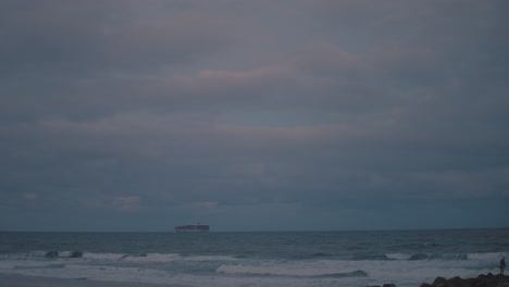 a large cargo ship sails by on the horizon of an afternoon beach where the setting sun spills light colours of blue and pink into the dangerous, stormy sky as well as the dark and moody water below