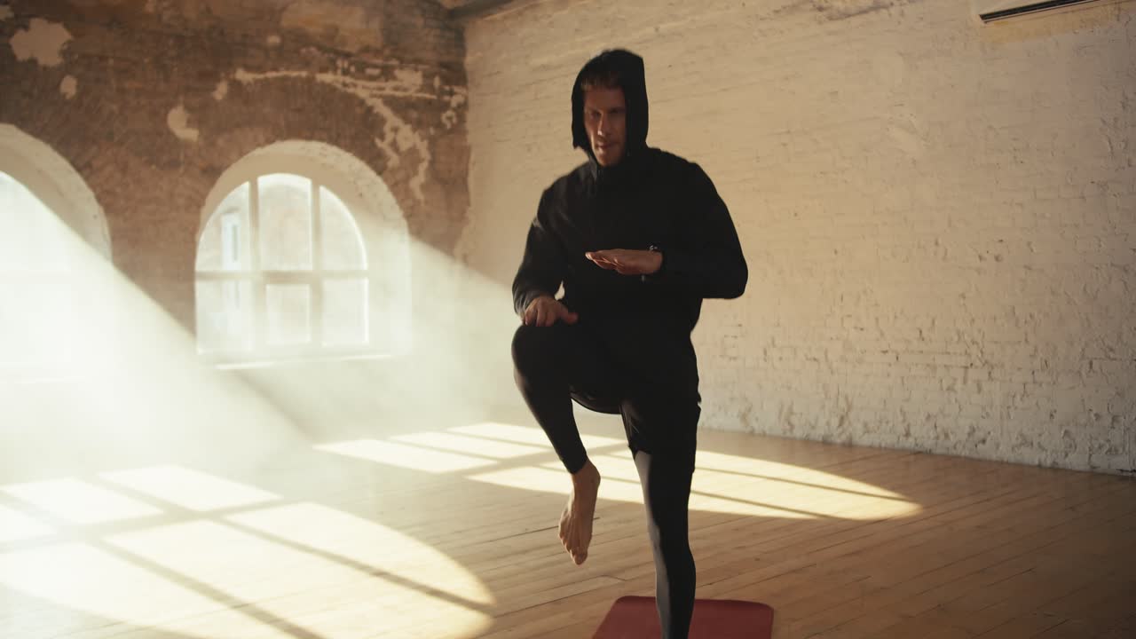A man in a black hooded sportswear warms up and runs in place on a special mat in a sunny brick hall
