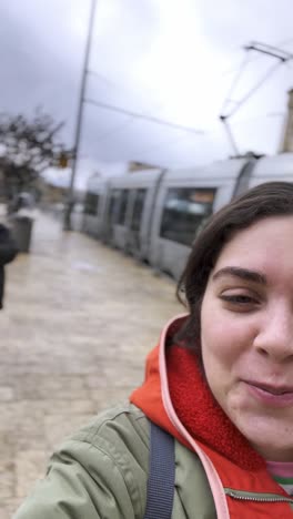 A woman is smiling and wearing a green jacket with an orange hood, taking video selfie.